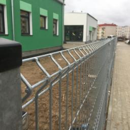 GERMAPLAN System Sp. z o.o. - Szare ogrodzenie systemowe z metalowych paneli na tle zielonego budynku i chodnika z kostki brukowej.