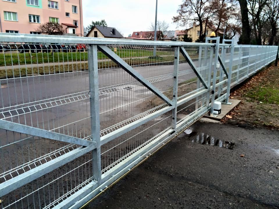 Przesuwna brama z metalowej siatki panelowej z automatyką, widok z zewnątrz na tle chodnika i budynków.