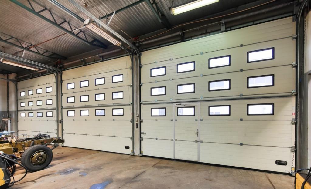 Dwie segmentowe bramy garażowe w kolorze kremowym z rzędami okienek, widoczne wewnątrz warsztatu, jedna z drzwiami wejściowymi wbudowanymi w bramę.