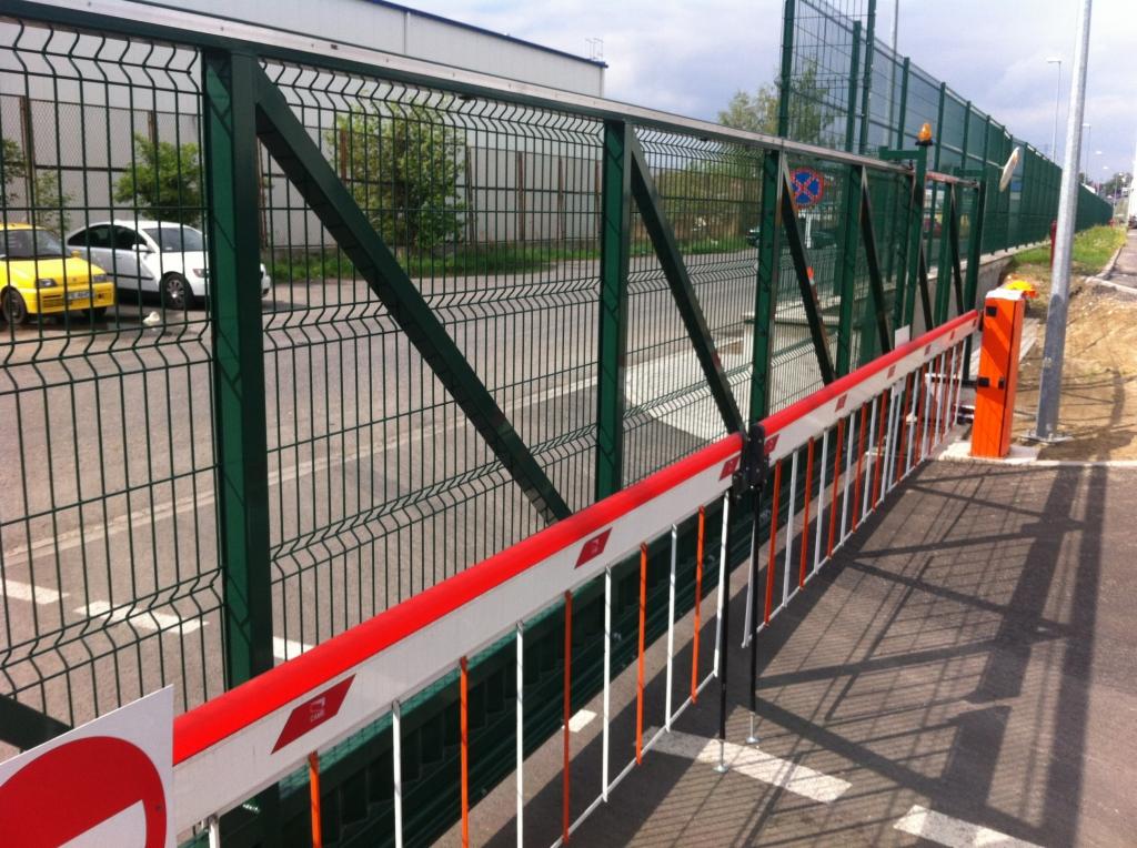 Szlaban z czerwono-białą belką zaporową, zamontowany przy zielonym ogrodzeniu panelowym z siatki, w tle budynek przemysłowy i zaparkowane samochody.