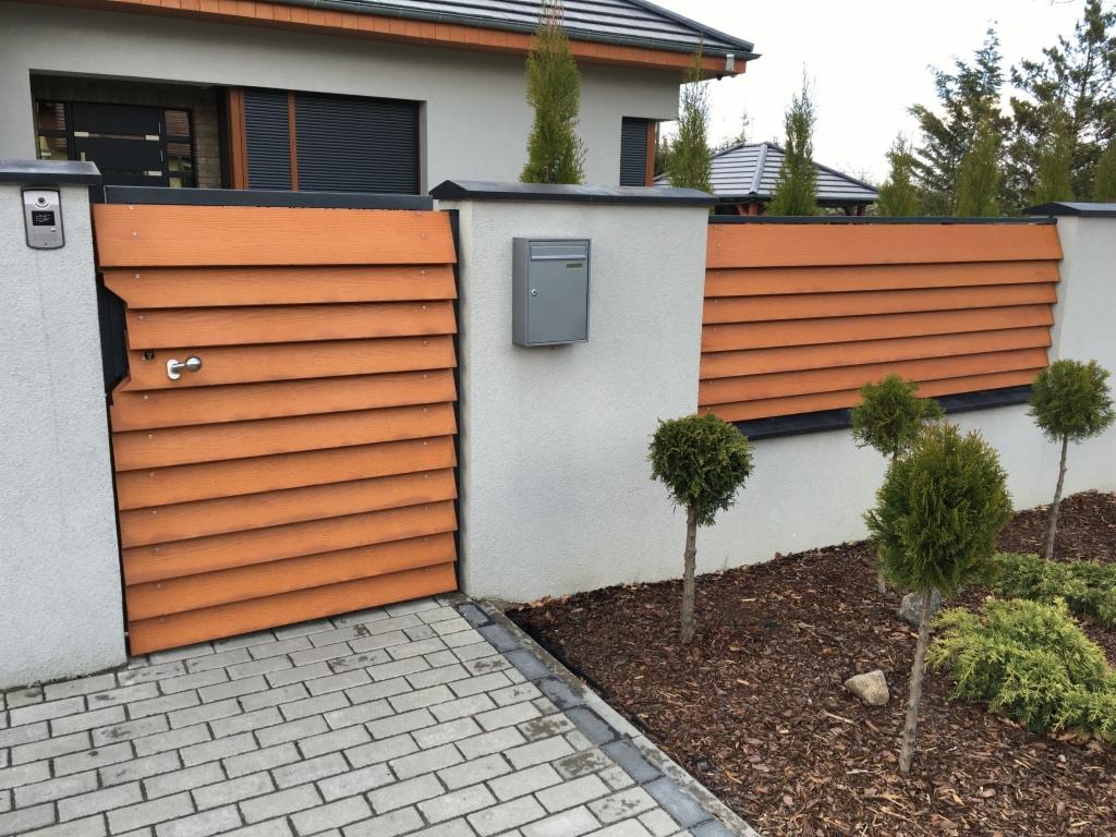 Nowoczesna furtka z poziomych desek kompozytowych w kolorze drewna, wkomponowana w ogrodzenie o podobnym designie, z widoczną skrzynką na listy i kostką brukową na podjeździe