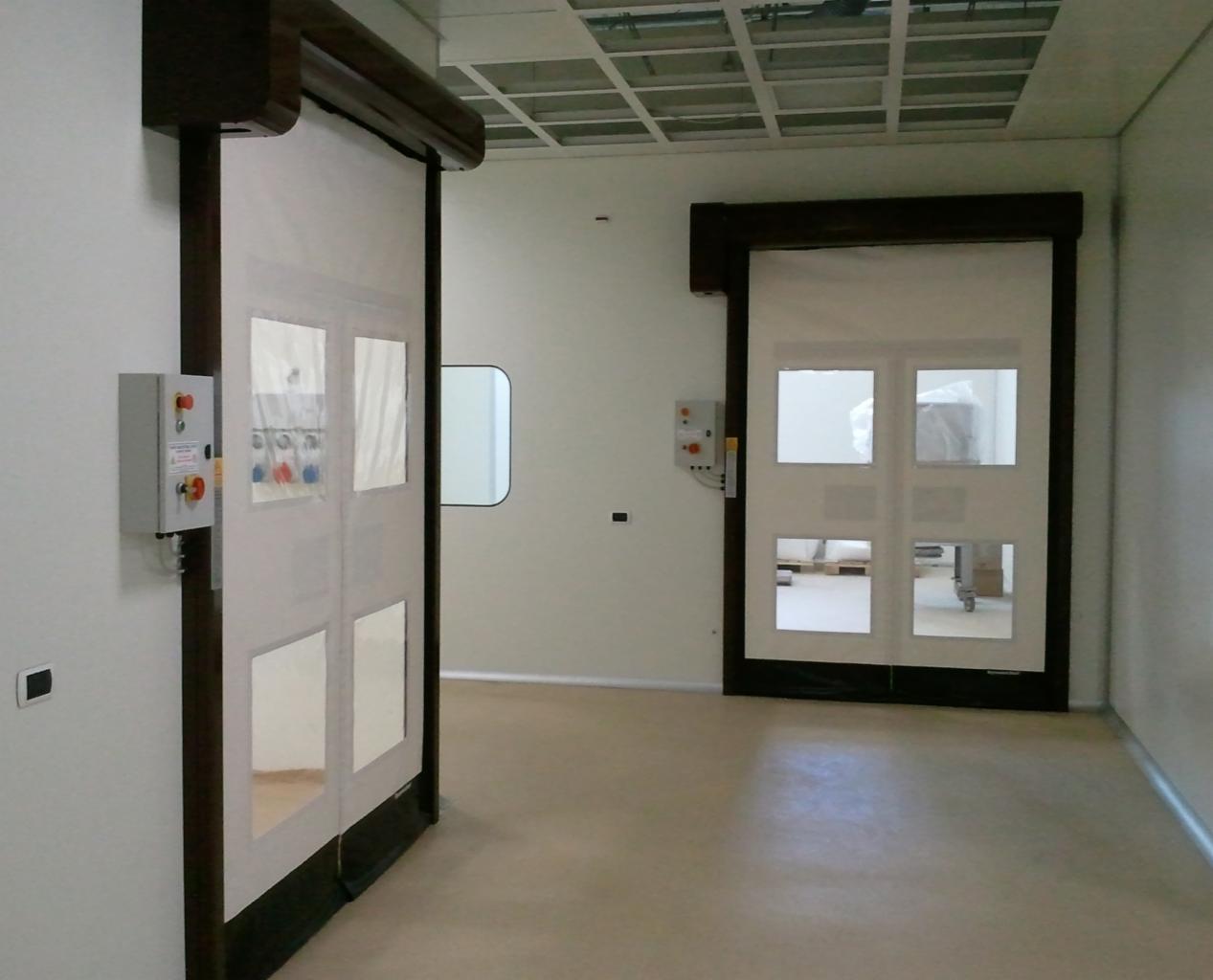 Dwie przesuwne bramy przemysłowe z brązowymi obramowaniami i przezroczystymi okienkami, zamontowane w sterylnym pomieszczeniu z szarą podłogą i panelowym sufitem.