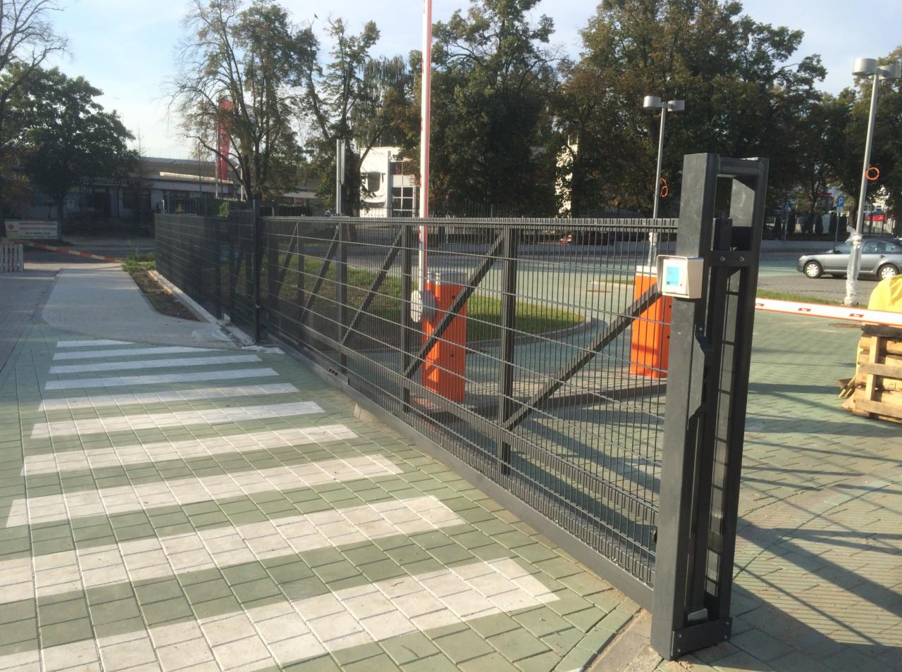 Przesuwna brama z siatki metalowej, z automatycznym szlabanem i panelem sterowania na słupku, widoczna na tle chodnika z kostki brukowej i budynków.