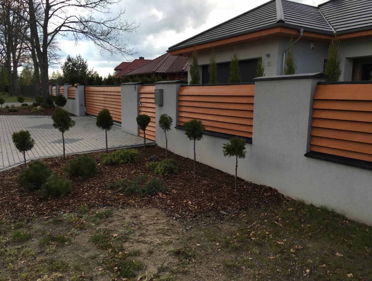 Nowoczesne ogrodzenie posesji z poziomych, drewnopodobnych paneli w kolorze ciepłego brązu, osadzone na szarych, murowanych słupkach. Wzdłuż ogrodzenia posadzone małe, kuliste drzewka i krzewy...