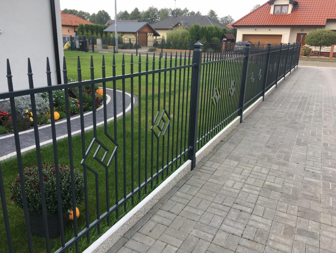 Ciemnoszare, metalowe ogrodzenie z geometrycznym wzorem rombu, oddzielające posesję z trawnikiem i rabatami kwiatowymi od kostki brukowej podjazdu.