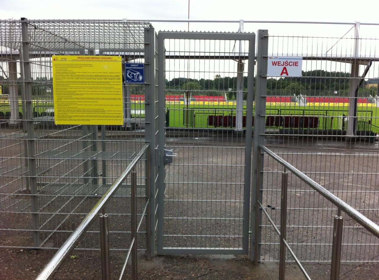 Otwarta metalowa furtka z siatki na stadion, widoczny napis 'Wejście A' oraz regulamin imprezy masowej, w tle trybuny stadionu i zieleń.