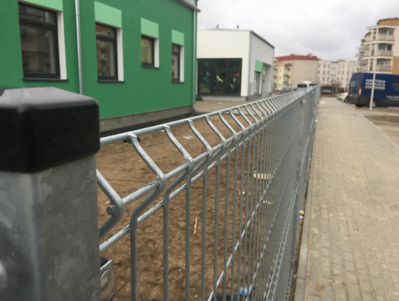 Szare ogrodzenie systemowe z metalowych paneli na tle zielonego budynku i chodnika z kostki brukowej.