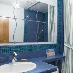 Niebiesko-zielona łazienka z mozaikową glazurą, umywalką nablatową, lustrem z metalową ramą i zasłoną prysznicową w paski.
