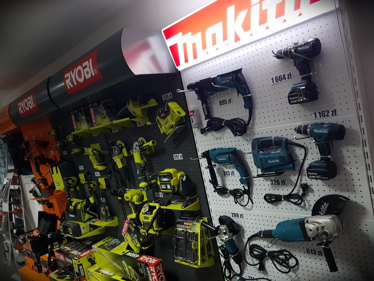 Ekspozycja elektronarzędzi marek Ryobi i Makita w sklepie, prezentująca wiertarki, szlifierki kątowe i piły, z widocznymi cenami na białym perforowanym panelu.