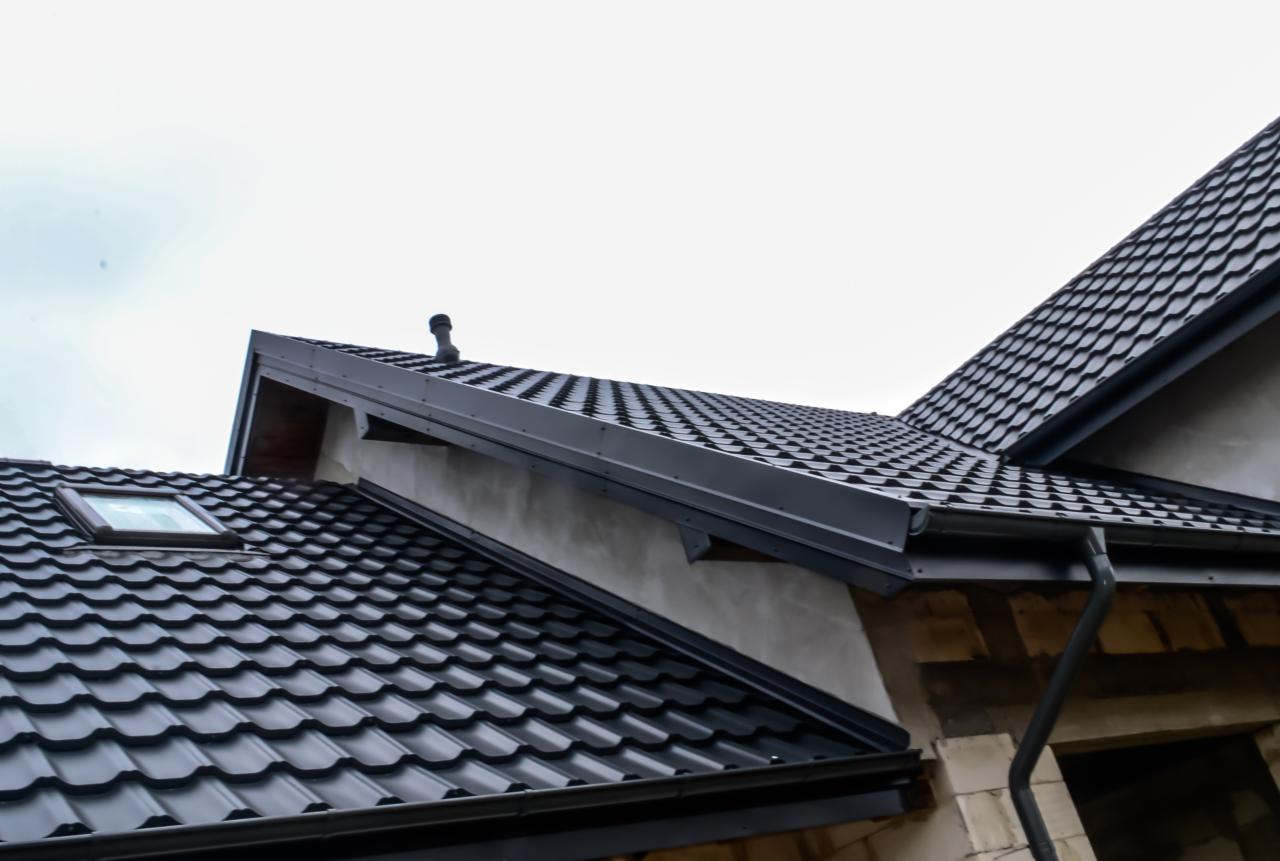 Fragment dachu z czarną blachodachówką modułową, lukarną i orynnowaniem, widoczny kominek wentylacyjny i surowa elewacja budynku w tle.