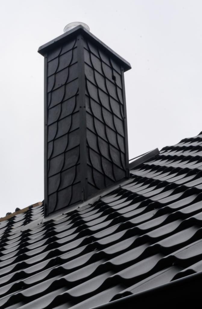 Czarny komin obłożony łupkiem na tle czarnego dachu z blachy trapezowej, widoczne połączenie komina z dachem.