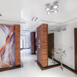 Nowoczesny korytarz z białymi, błyszczącymi płytkami podłogowymi, ceglanymi filarami, lustrem i przesuwnymi drzwiami z abstrakcyjnym wzorem w odcieniach fioletu i pomarańczy.