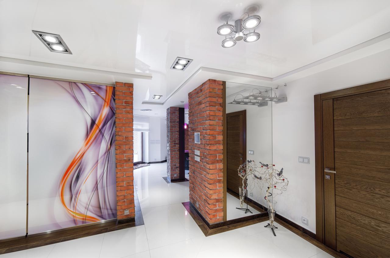 Nowoczesny korytarz z białymi, błyszczącymi płytkami podłogowymi, ceglanymi filarami, lustrem i przesuwnymi drzwiami z abstrakcyjnym wzorem w odcieniach fioletu i pomarańczy.