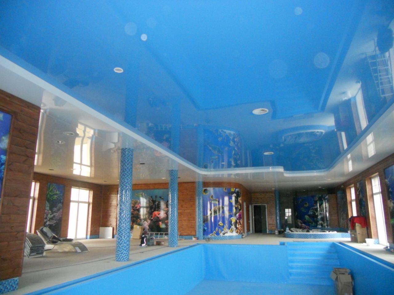 Wnętrze basenu w trakcie wykończenia, z błękitnym, błyszczącym sufitem podwieszanym imitującym wodę, drewnianymi ścianami z motywami morskimi i kolumnami wykończonymi mozaiką.