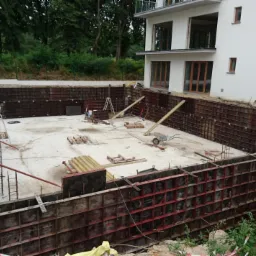 Głęboki wykop pod fundamenty domu, zabezpieczony szalunkiem, z widocznym znakiem ostrzegawczym 'Uwaga: Głębokie wykopy', na tle białego budynku w stanie surowym otoczonego zielenią.