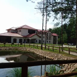 Dwukondygnacyjny budynek z czerwonym dachem, widoczny za stawem z kamiennym brzegiem i drewnianym ogrodzeniem, otoczony drzewami.