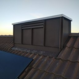 PS Dach - Brązowy komin wentylacyjny z metalowym wykończeniem na brązowym dachu z dachówki ceramicznej, widoczny panel solarny. Ujęcie z dołu, błękitne niebo w tle.