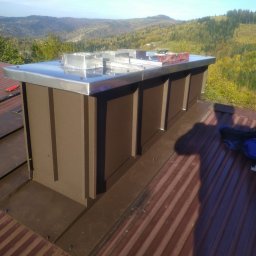 PS Dach - Nowa obudowa komina z brązowego metalu i błyszczącej blachy na dachu z widokiem na zalesione wzgórza w oddali, poziomica na górze.