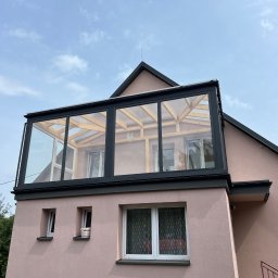 PS Dach - Różowy dom z dobudowaną przeszkloną werandą na dachu, czarne obramowanie, jasne niebo w tle. Widok z dołu, na ścianie widoczne okna.