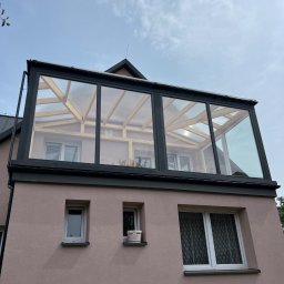 PS Dach - Szklana nadbudowa dachu z widoczną drewnianą konstrukcją, zamontowana na różowym budynku z oknami. Widok z dołu, pochmurne niebo w tle.