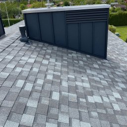 PS Dach - Dach pokryty szarym gontem bitumicznym z widocznym kominem wentylacyjnym i obróbkami blacharskimi w kolorze grafitowym na tle zieleni.