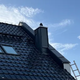 Ciemna dachówka ceramiczna z oknem dachowym i nowoczesnym kominem na tle błękitnego nieba z chmurami. Widoczna drabina i rusztowanie.