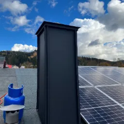 Dach pokryty papą z czarnym kominem, panele słoneczne i butla gazowa na tle błękitnego nieba z chmurami. Widok z góry.