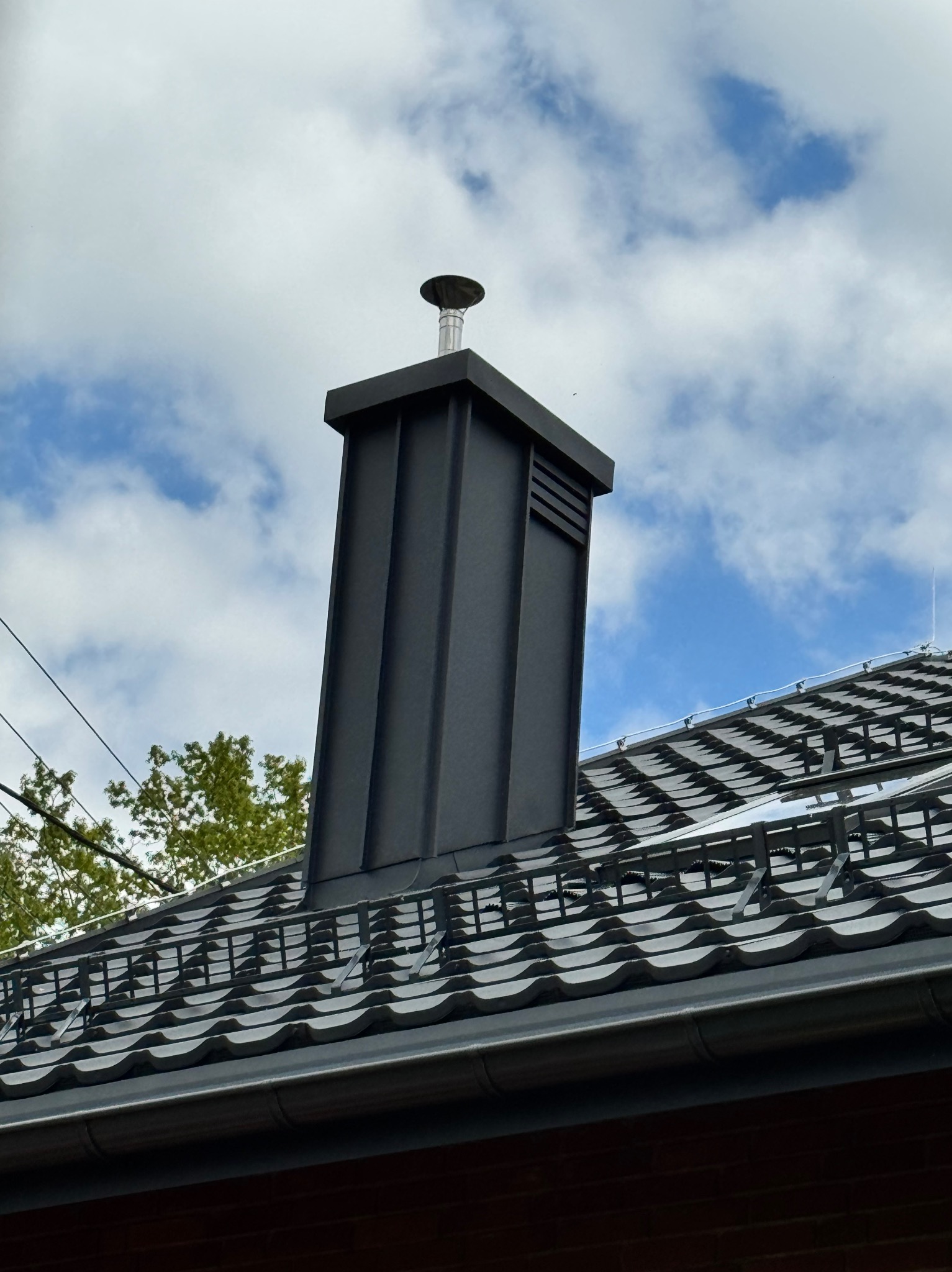 Nowoczesny, grafitowy komin z blachy na dachu z dachówki ceramicznej w Wiśle, na tle błękitnego nieba z chmurami. Widoczne zabezpieczenia przeciwśniegowe.