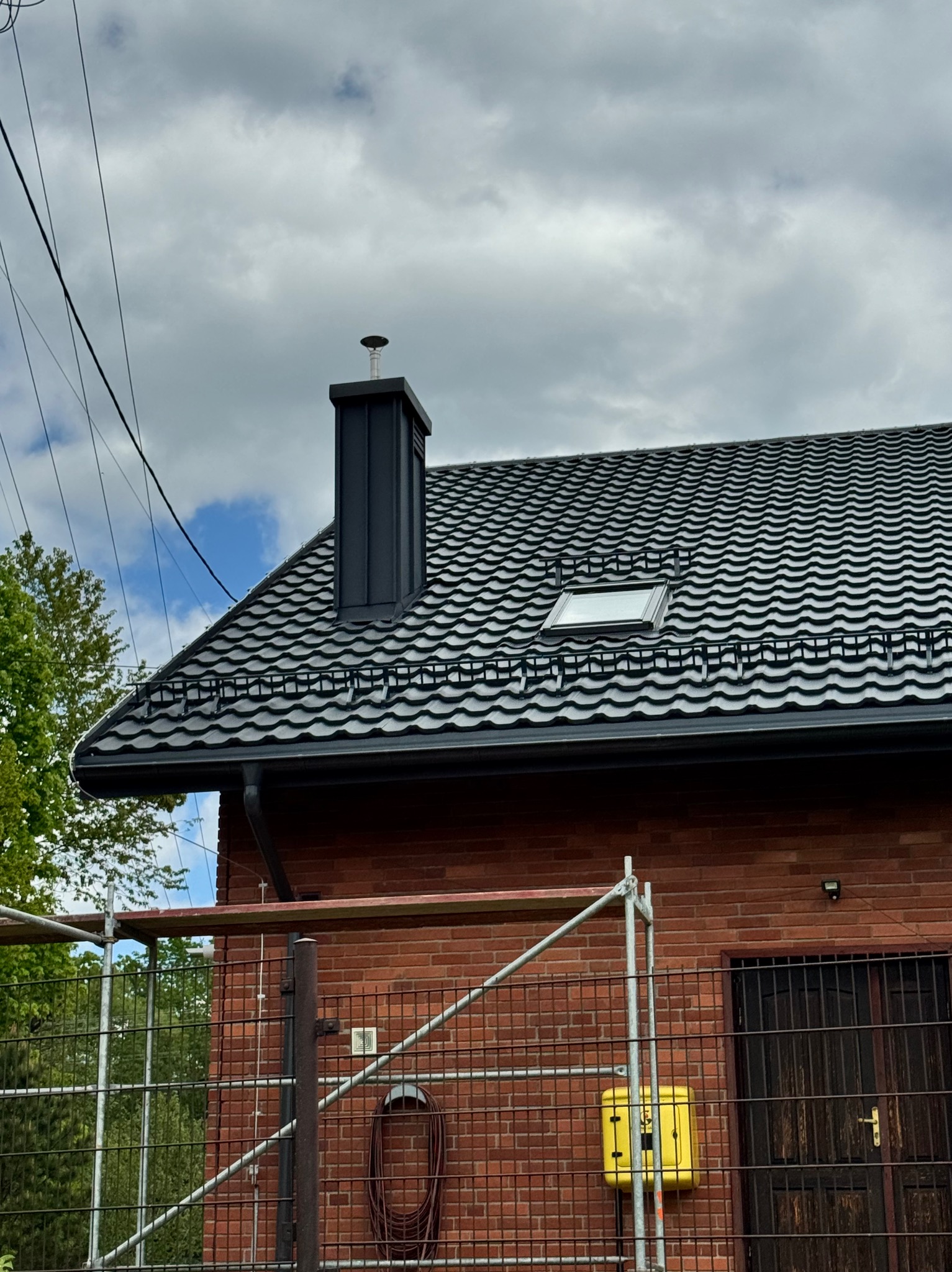 Dom z nowym, ciemnym dachem z dachówki falistej i czarnym kominem. Widoczne rusztowanie i okno dachowe. Ujęcie z dołu, pochmurne niebo w tle.