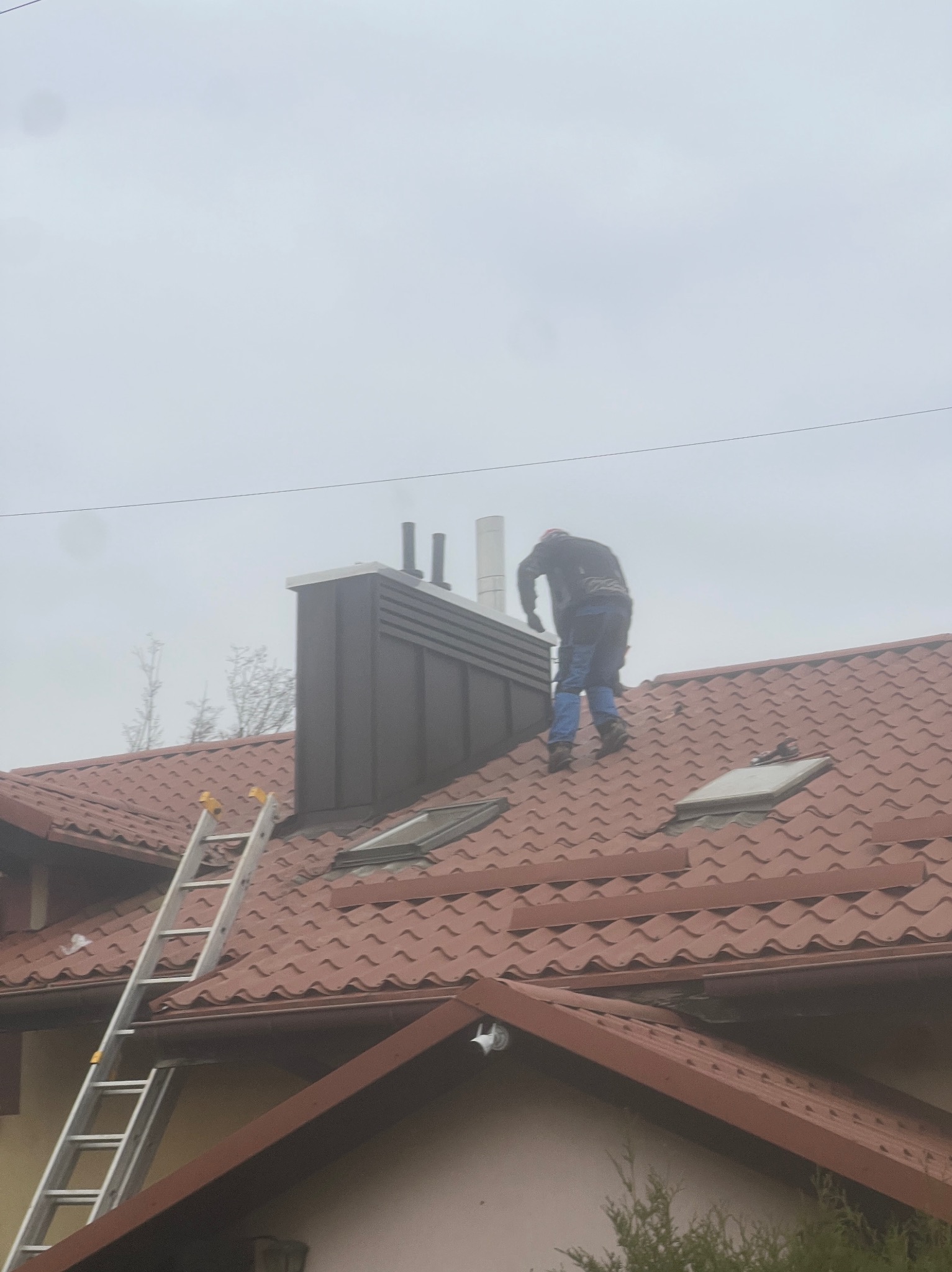 Praca na dachu: dekarz przy kominie z drabiny na tle pochmurnego nieba. Czerwona dachówka, okna dachowe. Usługi dekarskie.