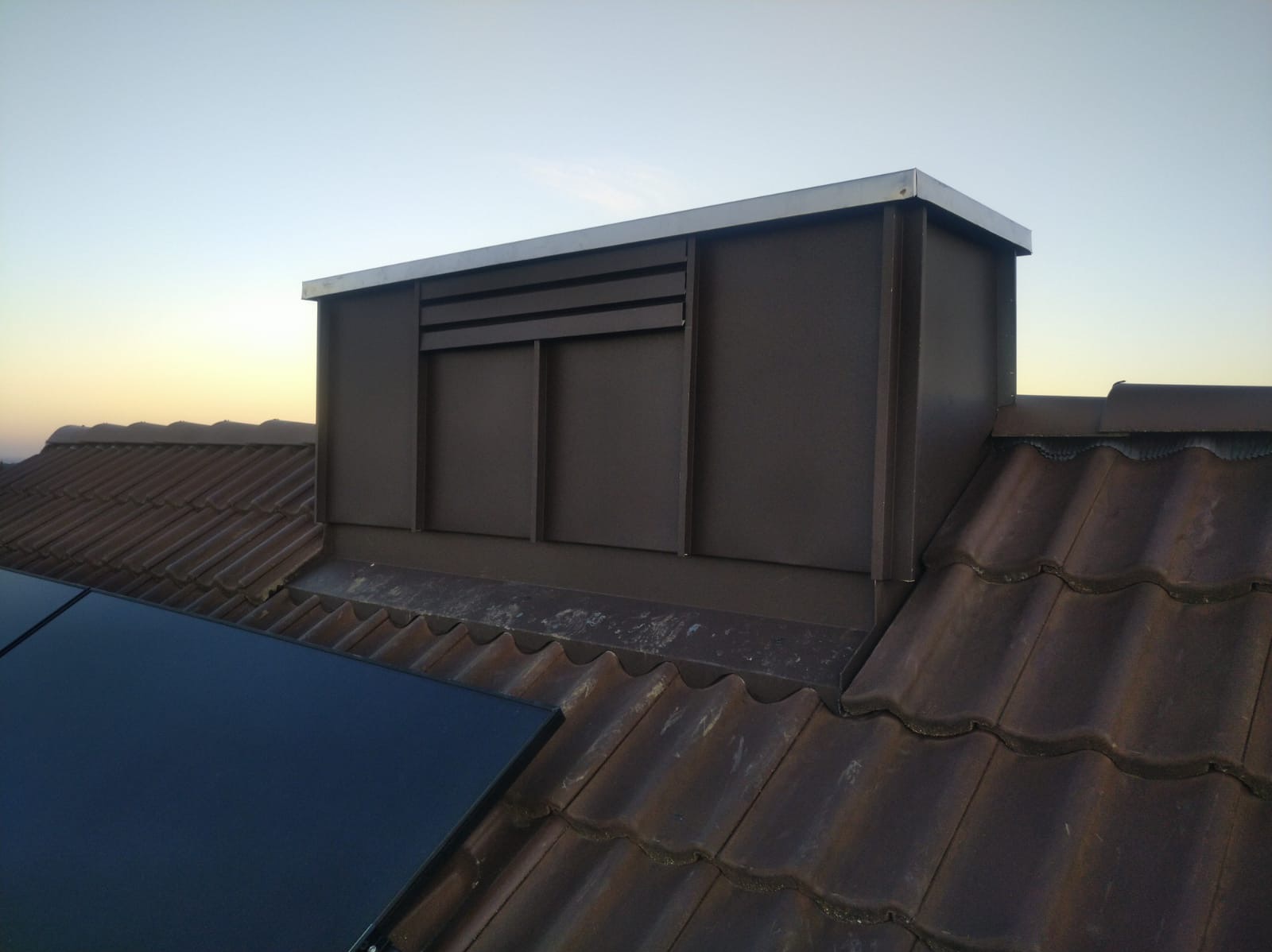 Brązowy komin wentylacyjny z metalowym wykończeniem na brązowym dachu z dachówki ceramicznej, widoczny panel solarny. Ujęcie z dołu, błękitne niebo w tle.