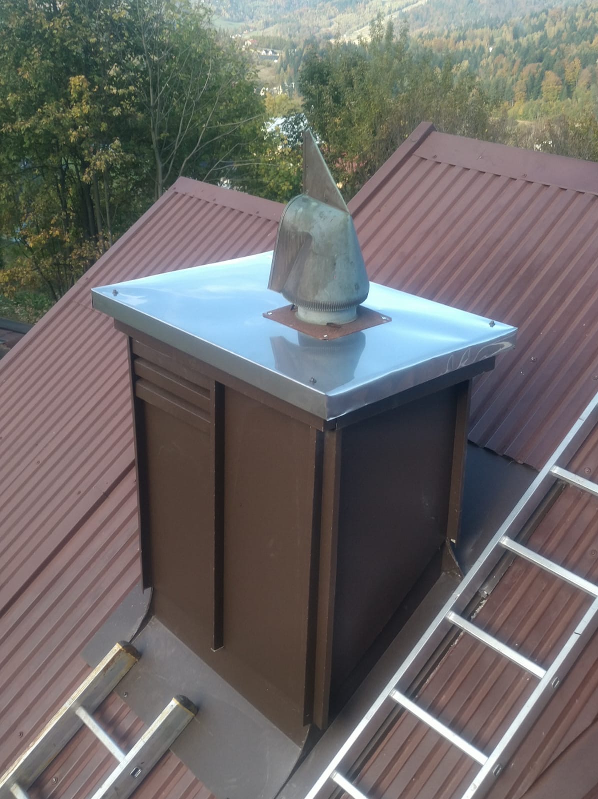 Nowy, brązowy komin z metalowym daszkiem i ozdobną, miedzianą końcówką na dachu z blachy trapezowej. W tle widok na zalesione wzgórza i drabina dekarska.