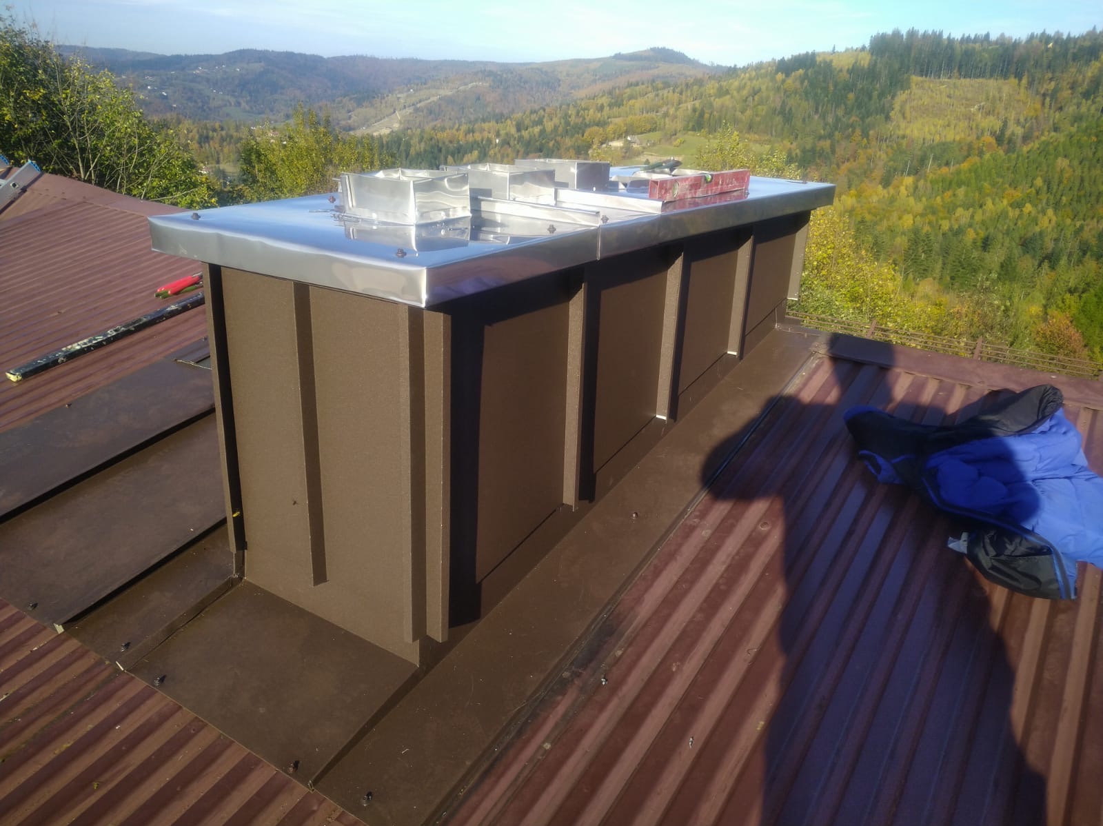 Nowa obudowa komina z brązowego metalu i błyszczącej blachy na dachu z widokiem na zalesione wzgórza w oddali, poziomica na górze.