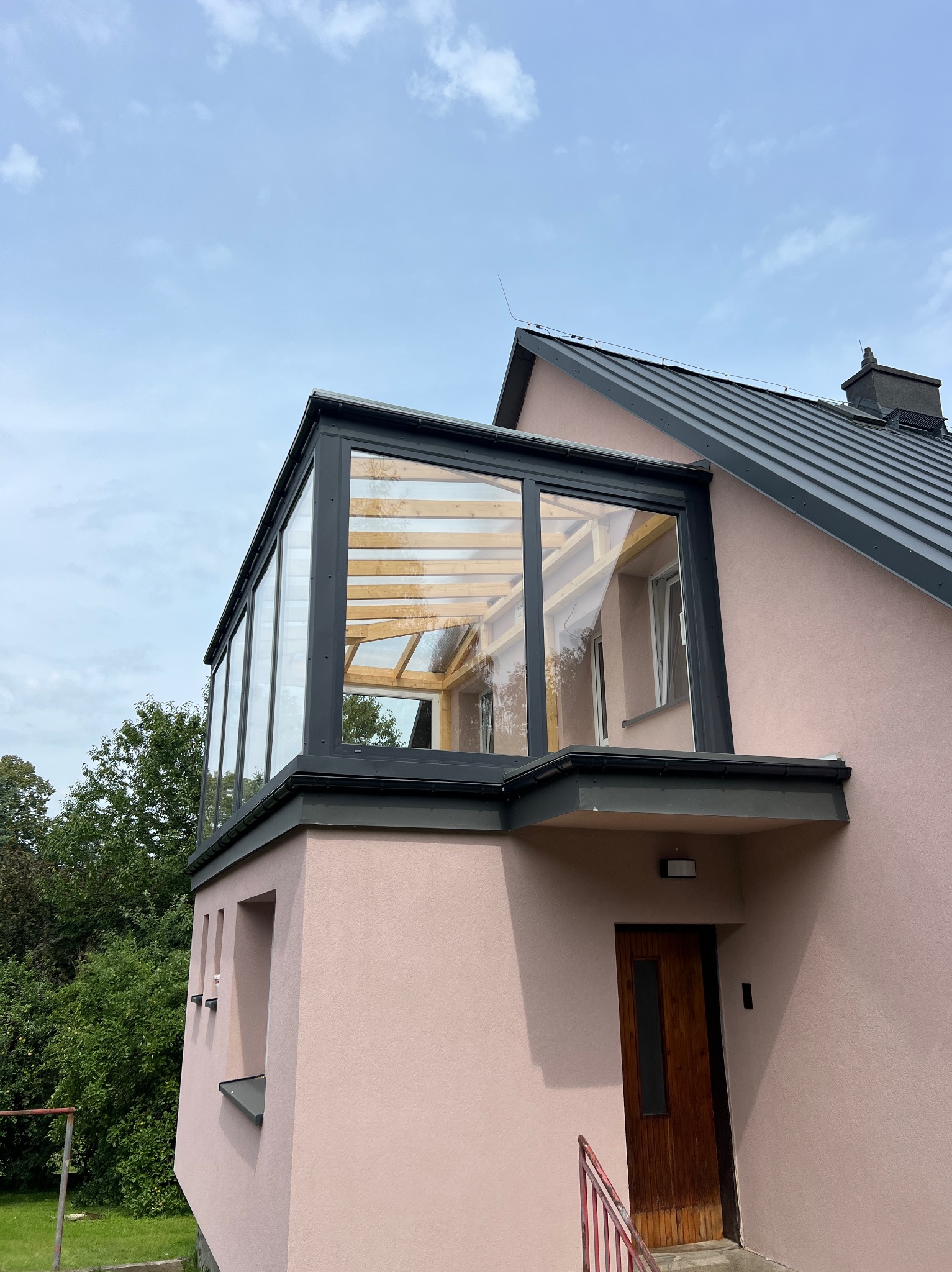 Dom z przeszkloną werandą na dachu, widok z dołu na jasnoróżową elewację i szary dach. Widoczne elementy obróbki blacharskiej i drewniana konstrukcja werandy.