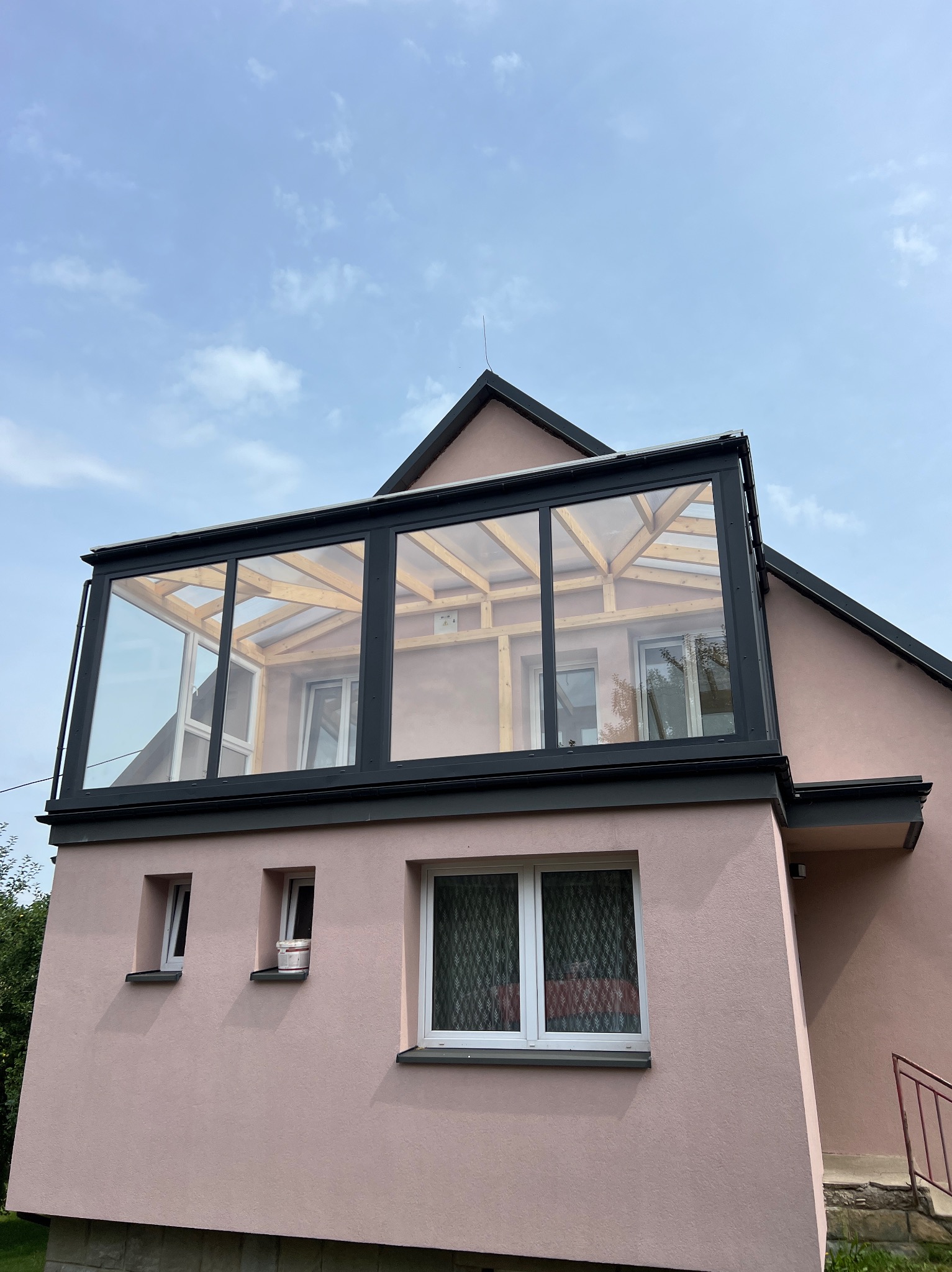 Różowy dom z dobudowaną przeszkloną werandą na dachu, czarne obramowanie, jasne niebo w tle. Widok z dołu, na ścianie widoczne okna.