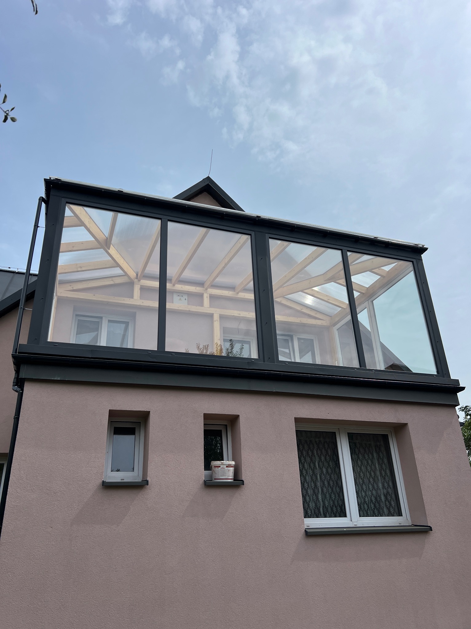 Szklana nadbudowa dachu z widoczną drewnianą konstrukcją, zamontowana na różowym budynku z oknami. Widok z dołu, pochmurne niebo w tle.