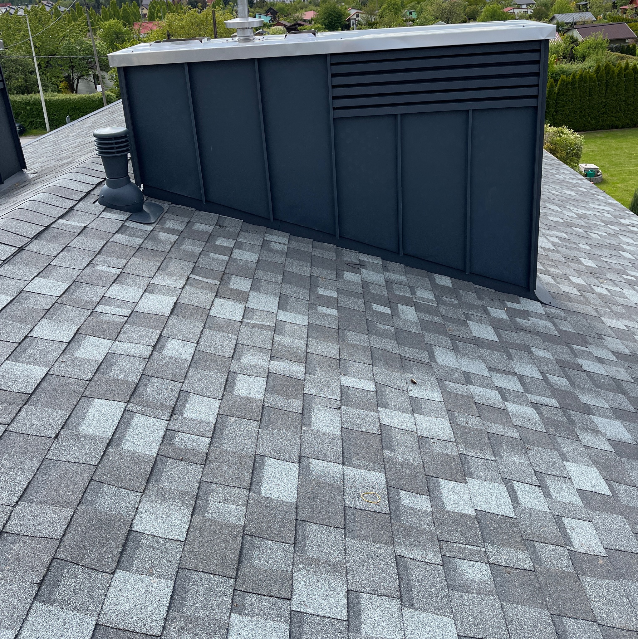 Dach pokryty szarym gontem bitumicznym z widocznym kominem wentylacyjnym i obróbkami blacharskimi w kolorze grafitowym na tle zieleni.