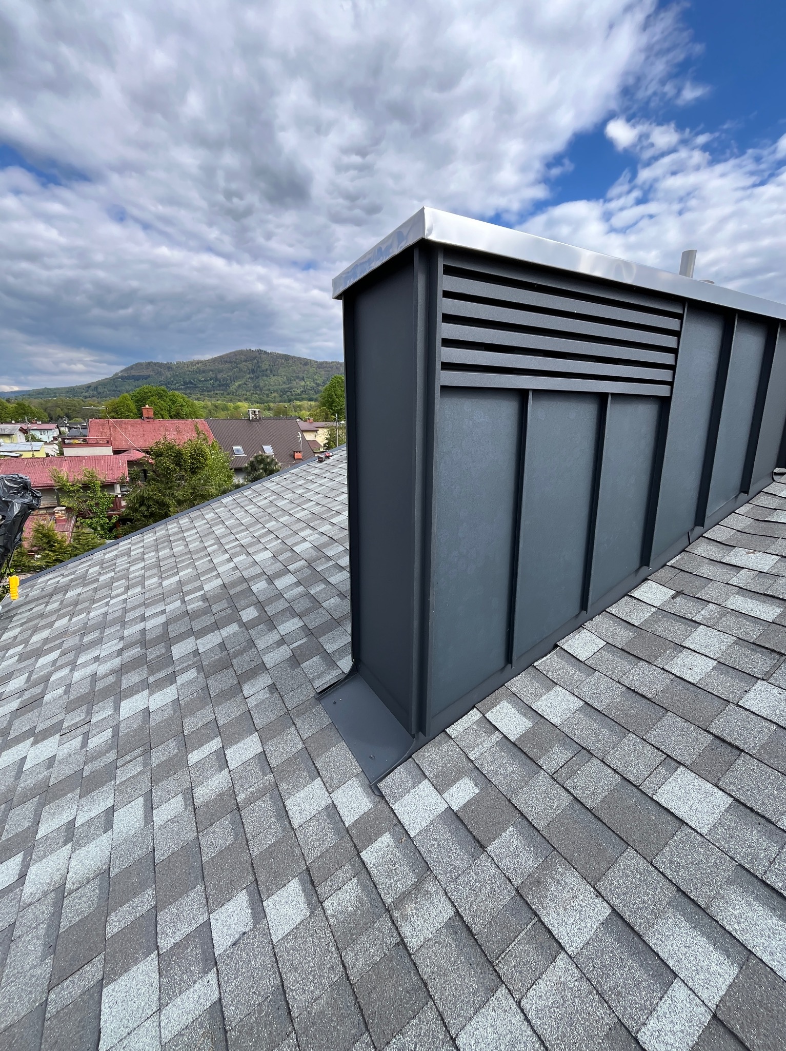 Szary dach pokryty gontem bitumicznym, z kominem wentylacyjnym i widokiem na góry w tle. Dach po remoncie, z bliska.