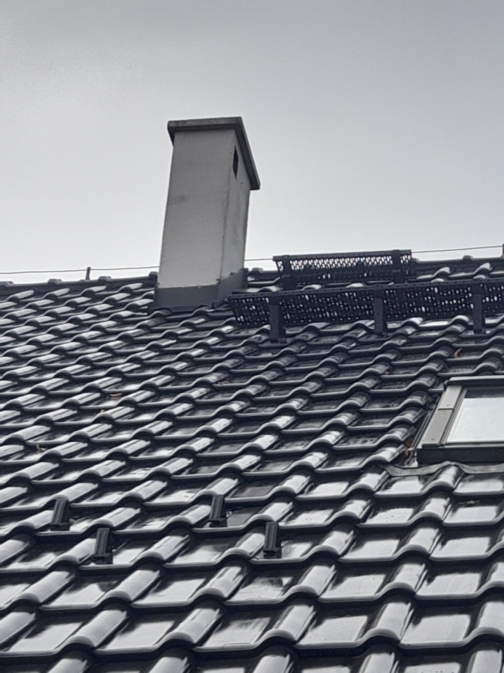 Błyszczący, czarny dach z dachówki ceramicznej z zamontowanymi stopniami kominiarskimi i kominem na tle pochmurnego nieba.