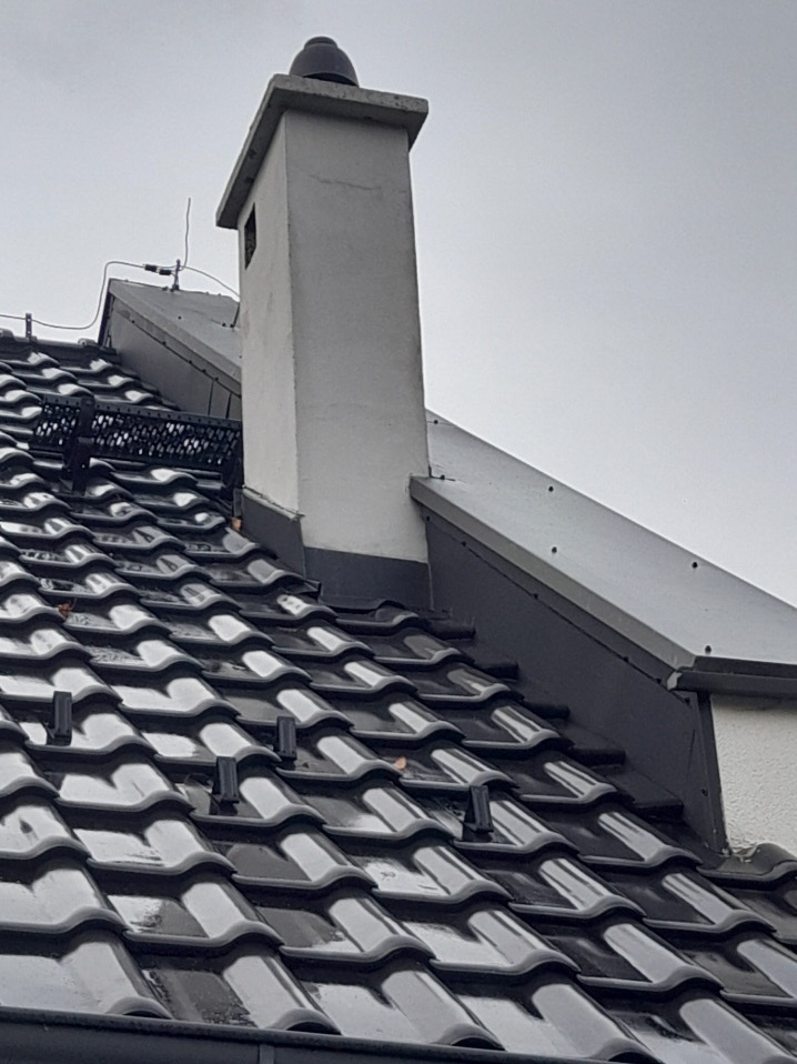 Błyszczący, ciemny dach ceramiczny z kominem w tle, zabezpieczony przed osuwaniem się śniegu. Widoczne obróbki blacharskie przy kominie.