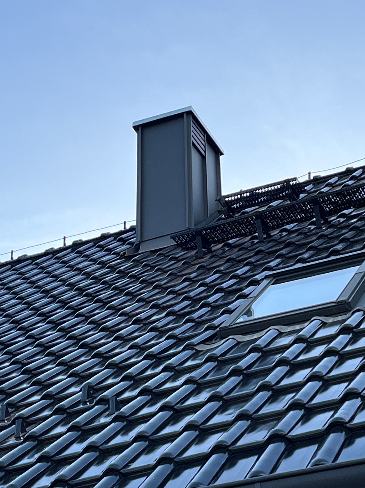 Błyszczący, czarny dach z dachówki ceramicznej z oknem dachowym i metalowym kominem wentylacyjnym na tle jasnego nieba. Widoczne zabezpieczenia przeciwśniegowe.