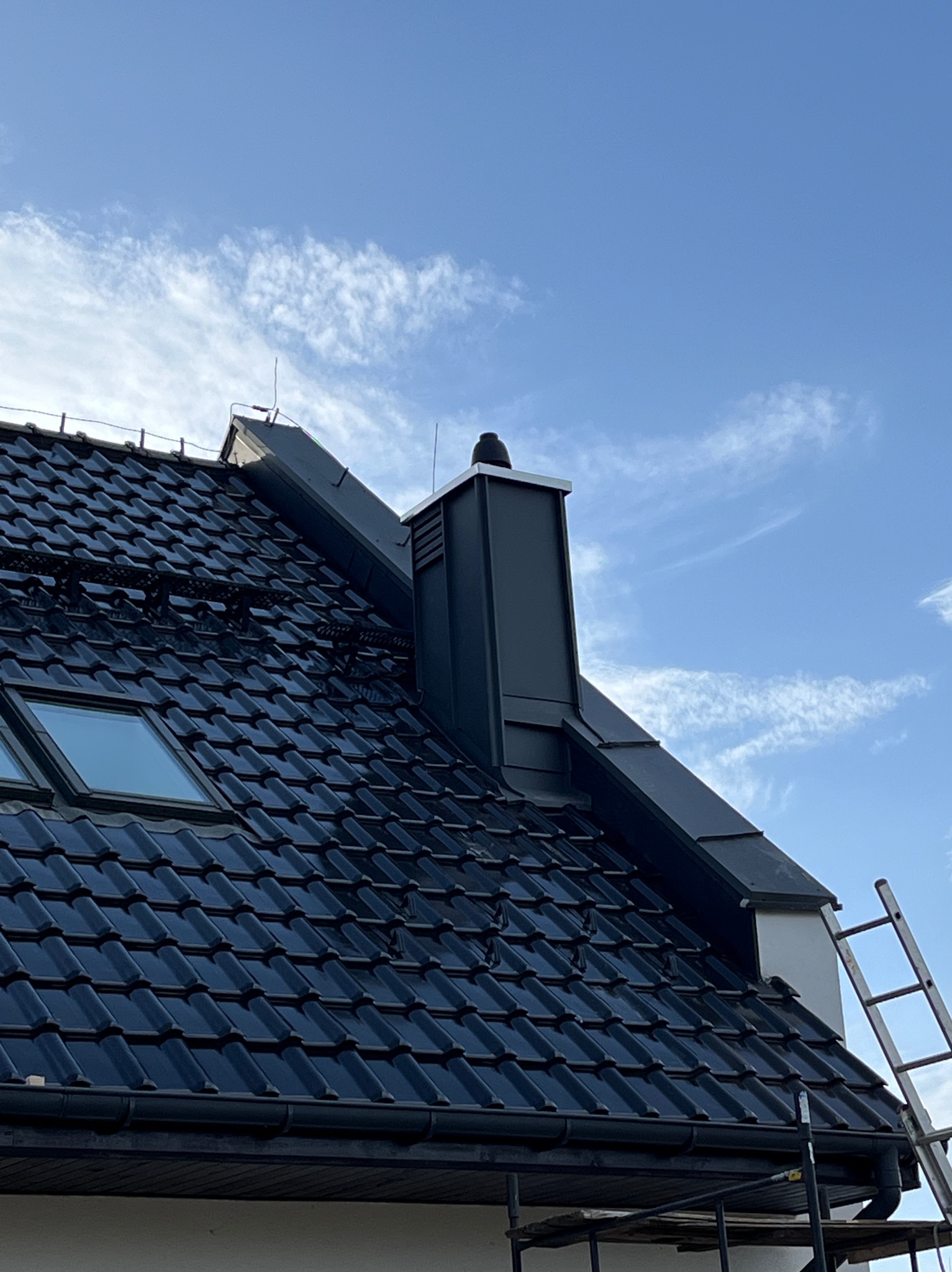 Ciemna dachówka ceramiczna z oknem dachowym i nowoczesnym kominem na tle błękitnego nieba z chmurami. Widoczna drabina i rusztowanie.