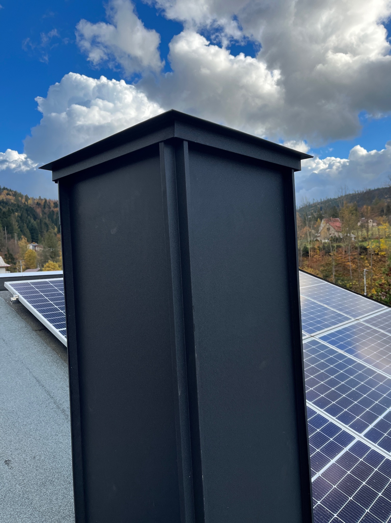 Czarny komin na dachu z panelami słonecznymi, błękitne niebo z chmurami w tle. Ujęcie z dołu podkreśla solidność konstrukcji i nowoczesne rozwiązania.