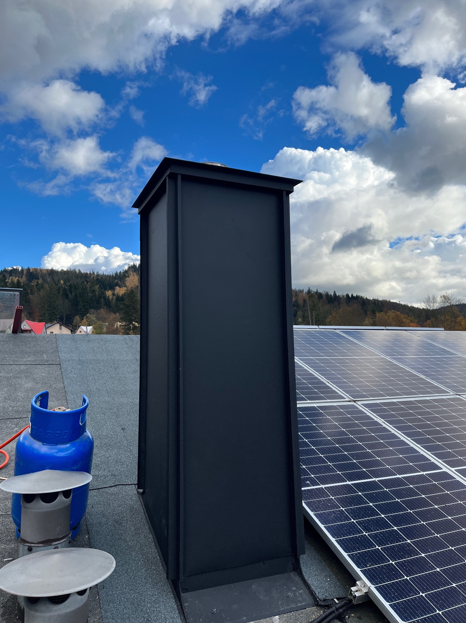 Dach pokryty papą z czarnym kominem, panele słoneczne i butla gazowa na tle błękitnego nieba z chmurami. Widok z góry.