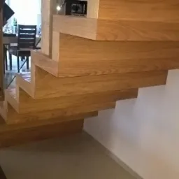 Detal drewnianych schodów wspornikowych z widocznym usłojeniem drewna dębowego, zamontowanych przy białej ścianie w nowoczesnym wnętrzu.