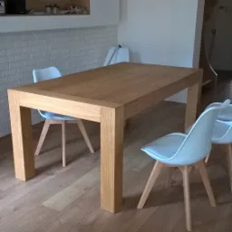 Jasny, prostokątny stół z litego drewna w nowoczesnej jadalni, otoczony białymi krzesłami na drewnianych nogach, z podłogą z paneli i białymi ścianami z cegieł oraz wnękami z półkami.