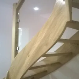 Klatka schodowa z jasnego drewna z zakrzywioną balustradą i szklanym panelem ochronnym, widok z dołu.