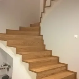 Drewniane schody z minimalistyczną, metalową balustradą w nowoczesnym domu. Widoczny fragment trójkątnego okna w ścianie. Pod schodami znajduje się schowek z odkurzaczem i butami.