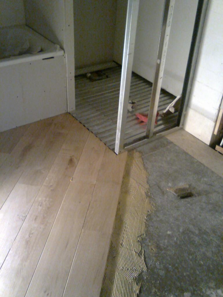 Montaż jasnych paneli podłogowych w łazience, widoczny klej na posadzce, metalowa konstrukcja kabiny prysznicowej i fragment wanny.