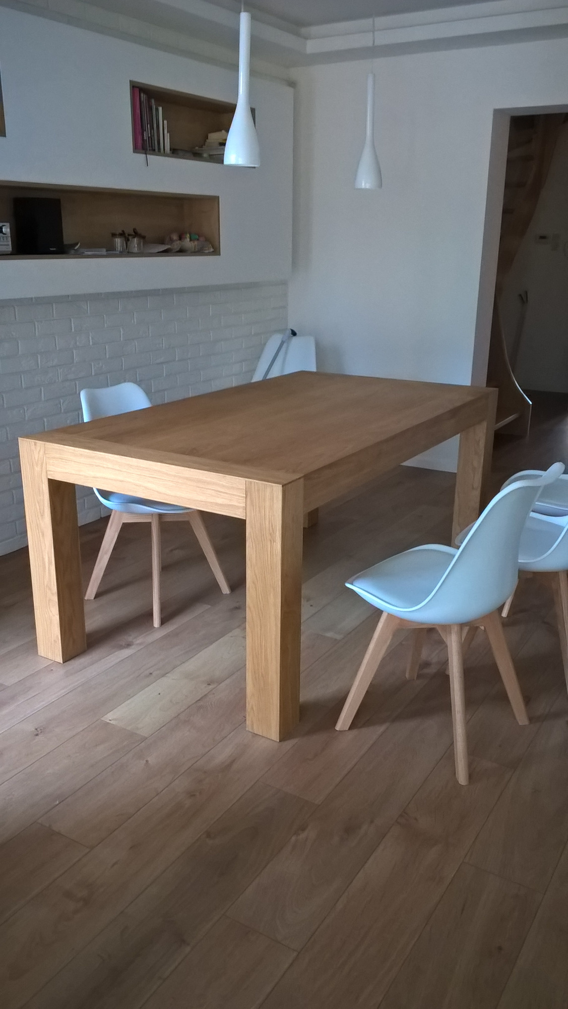 Jasny, prostokątny stół z litego drewna w nowoczesnej jadalni, otoczony białymi krzesłami na drewnianych nogach, z podłogą z paneli i białymi ścianami z cegieł oraz wnękami z półkami.